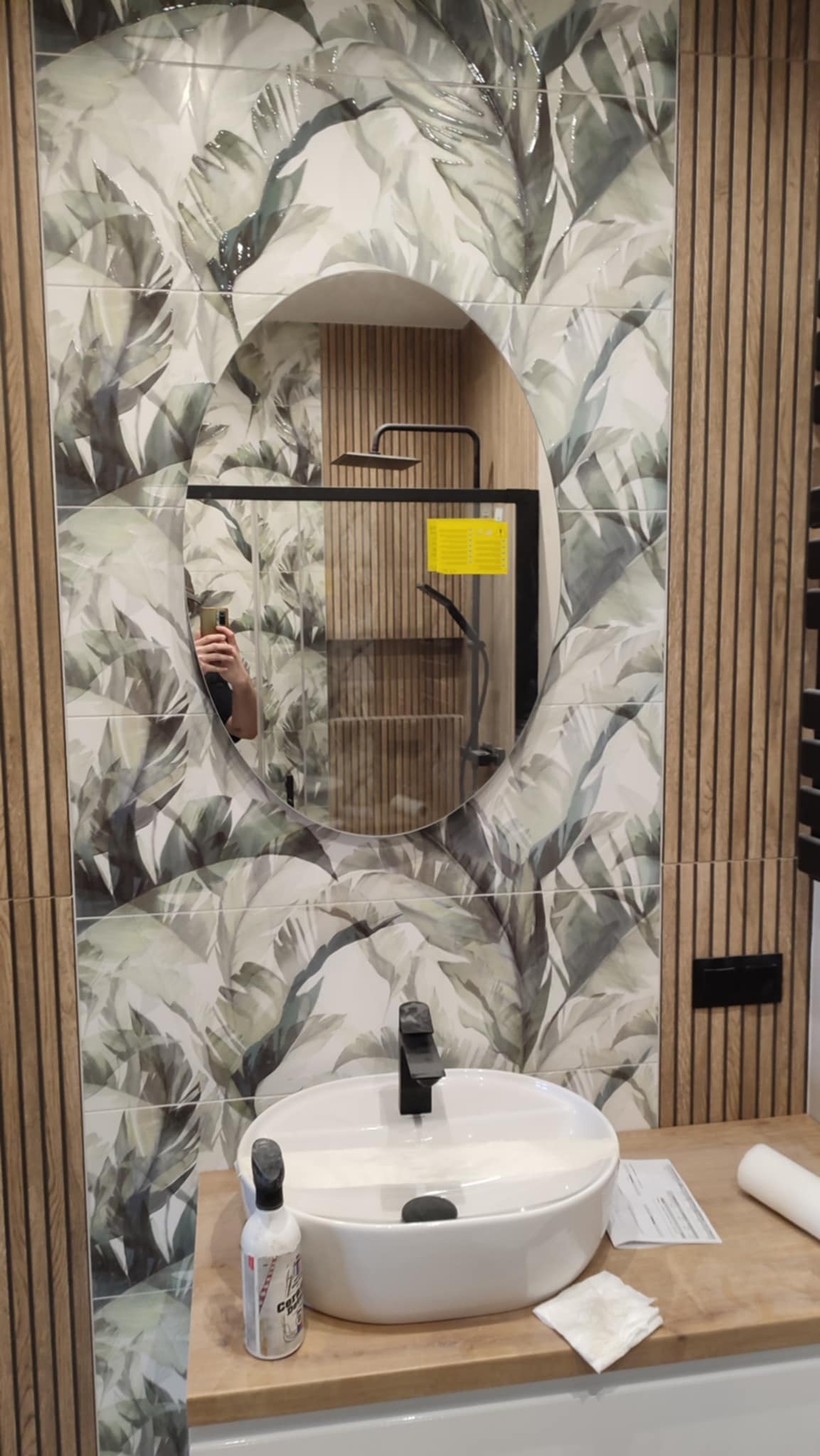 Wykończona łazienka w Koczergach z umywalką nablatową i lustrem w owalnej ramie. Na ścianie płytki z motywem liści i panele imitujące drewno.