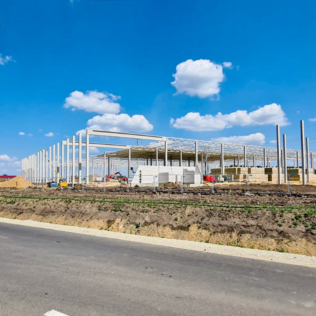 Konstrukcja stalowa hali w trakcie budowy, widok z zewnątrz, błękitne niebo z chmurami w tle, widoczne maszyny budowlane i materiały.