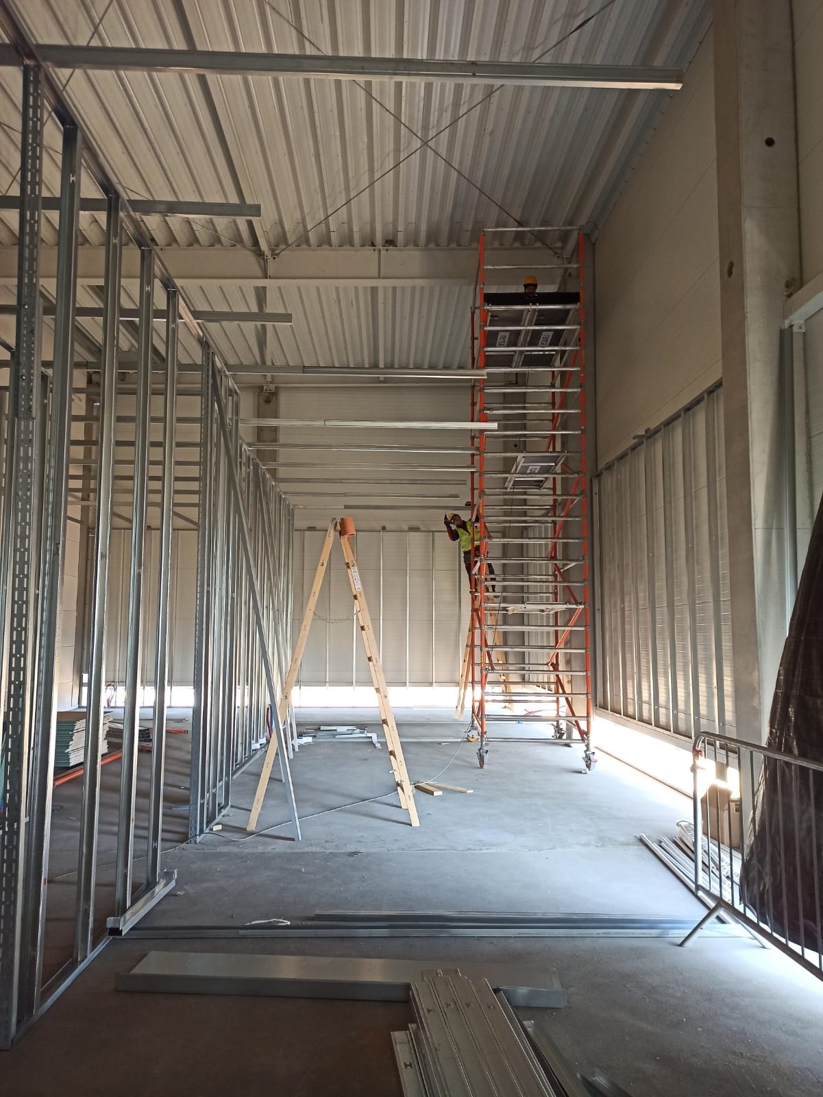 Montaż konstrukcji ścian działowych z profili stalowych w dużym pomieszczeniu. Widoczne dwie drabiny i pracownik na rusztowaniu. Metalowe profile na podłodze.