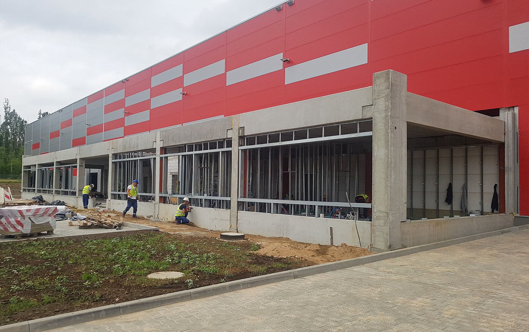 Budowa hali z czerwoną elewacją i stalową konstrukcją. Trzech pracowników w odblaskowych kamizelkach wykonuje prace montażowe na zewnątrz budynku.