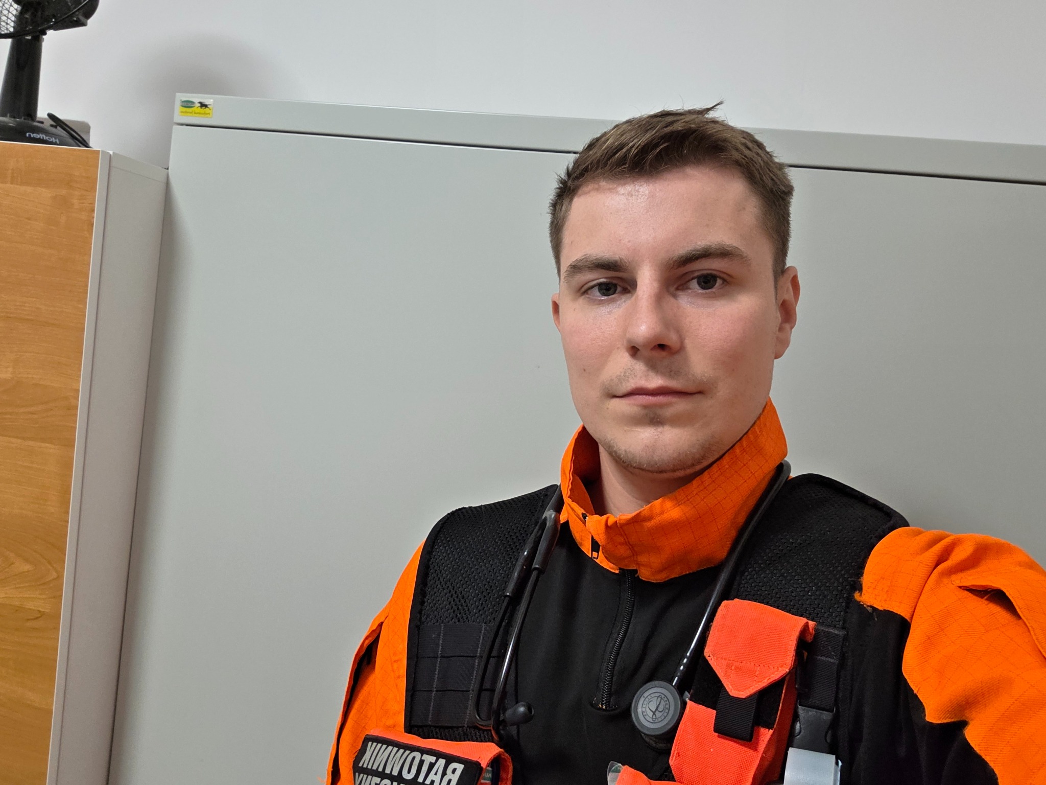 Młody mężczyzna w pomarańczowo-czarnym stroju ratownika medycznego ze stetoskopem na tle szarej szafki i wentylatora. Ujęcie z bliska, portret.