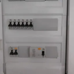 Biała skrzynka elektryczna z zainstalowanymi bezpiecznikami i wyłącznikami różnicowoprądowymi marki Ideal, widoczne oznaczenia CE. Instalacja wewnątrz budynku.