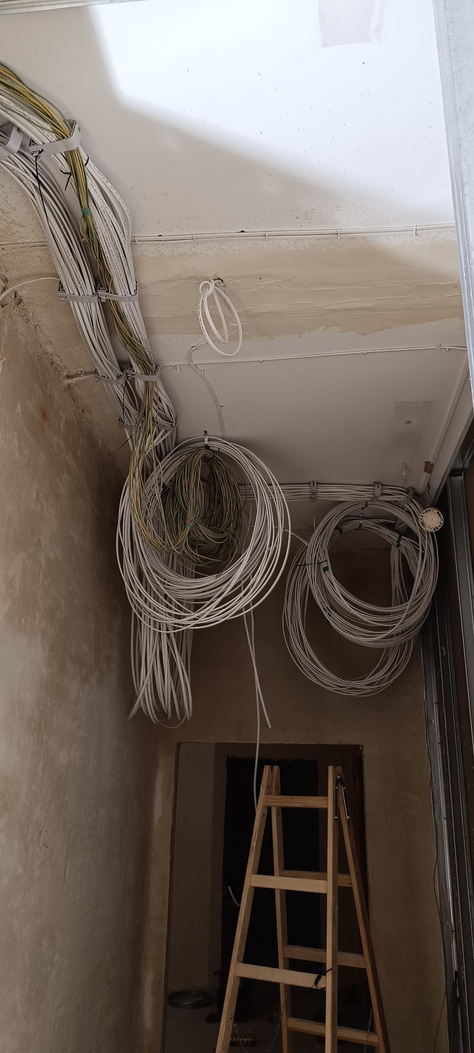 Wiązki kabli elektrycznych przymocowane do sufitu w surowym wnętrzu. Widoczna drabina malarska. Kable ułożone w okręgi. W tle otwór drzwiowy.