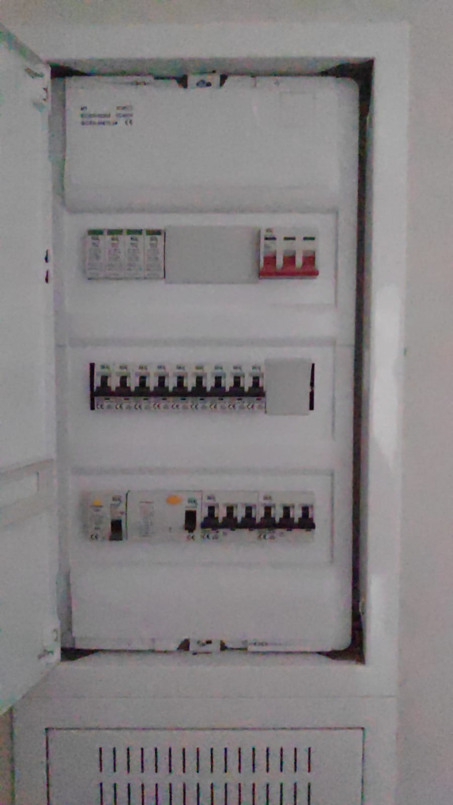 Otwarta skrzynka elektryczna z bezpiecznikami i wyłącznikami, widoczne zabezpieczenia różnicowoprądowe i nadprądowe w białej obudowie, zamontowana w ścianie.