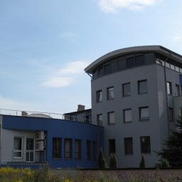 PPHU Iwbud - Nowoczesny budynek biurowy z szarymi ścianami i niebieskim akcentem, widoczne okna z ciemnymi ramami, niewielki balkon i zieleń w otoczeniu, błękitne niebo w tle.