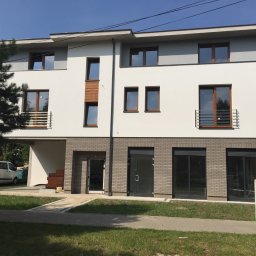 PPHU Iwbud - Nowoczesny budynek z brązowymi oknami PCV i szarą elewacją z cegły, jasne tło, dzień. Widoczne drzwi wejściowe i okna z barierkami.