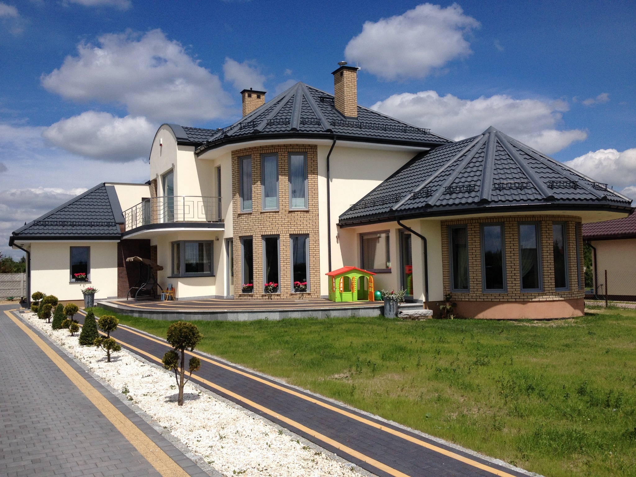 Elewacja dwukondygnacyjnego domu z szarą dachówką, beżowymi ścianami i ceglaną wstawką, otoczonego zielenią i ozdobnym chodnikiem z kostki brukowej.