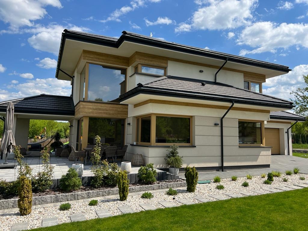 Elewacja domu jednorodzinnego z dużymi oknami i roletami wewnętrznymi, jasna fasada, widok z zewnątrz,  zagospodarowany teren zielony.