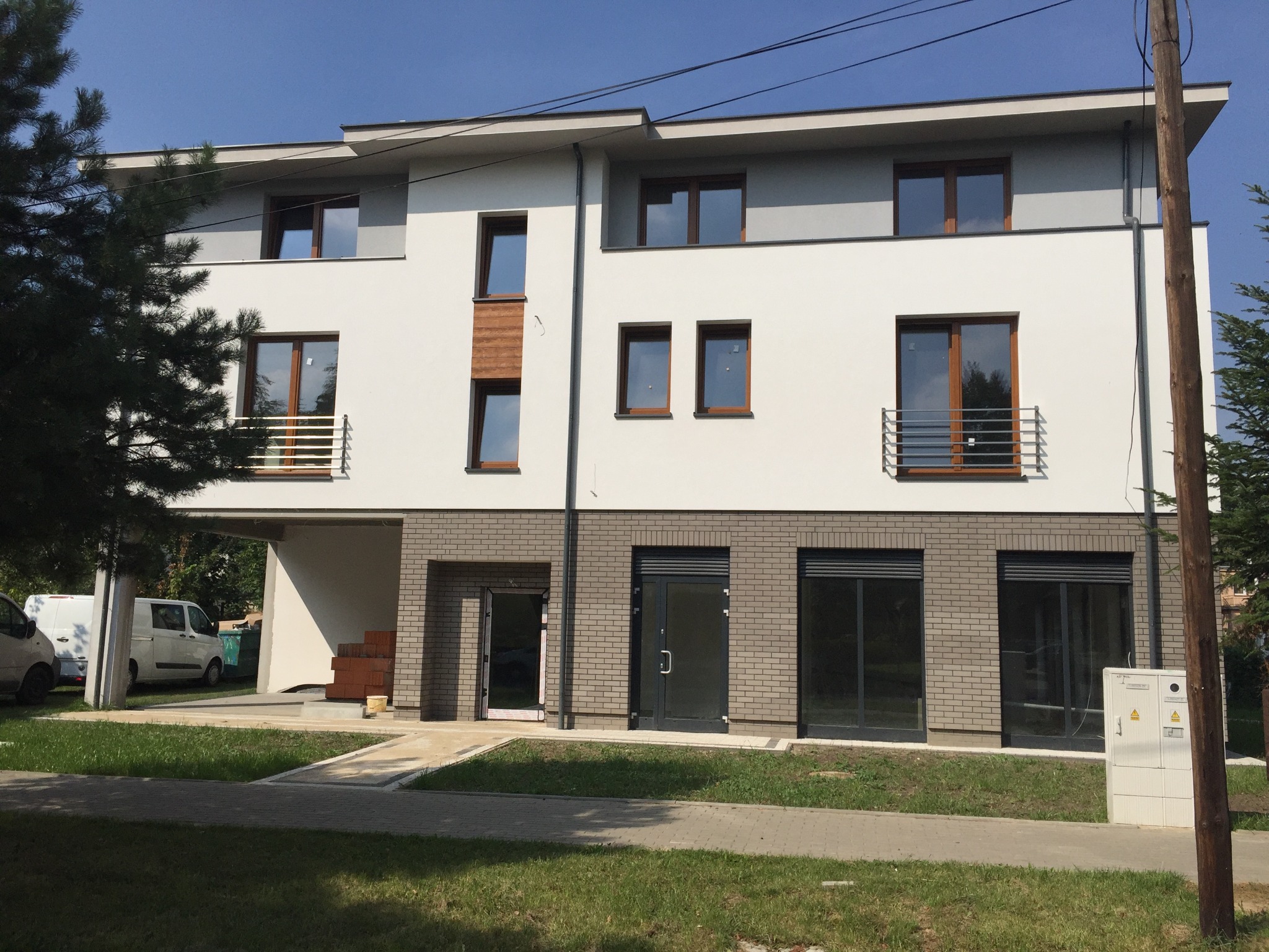 Nowoczesny budynek z brązowymi oknami PCV i szarą elewacją z cegły, jasne tło, dzień. Widoczne drzwi wejściowe i okna z barierkami.