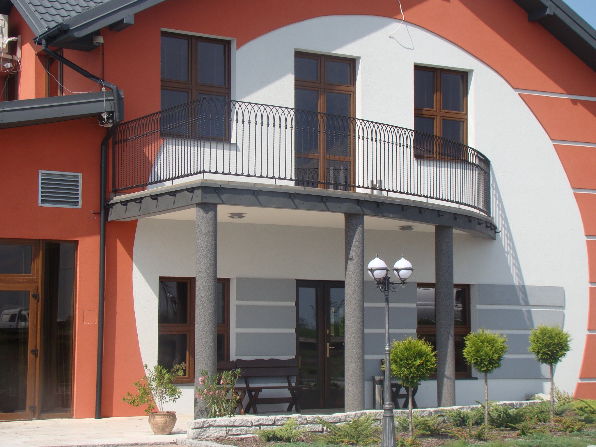 Elewacja domu z balkonem, okna z drewnianą ramą, dekoracyjne kolumny i lampa. Czerwono-biała kolorystyka, zadbany ogród z niskimi drzewkami.