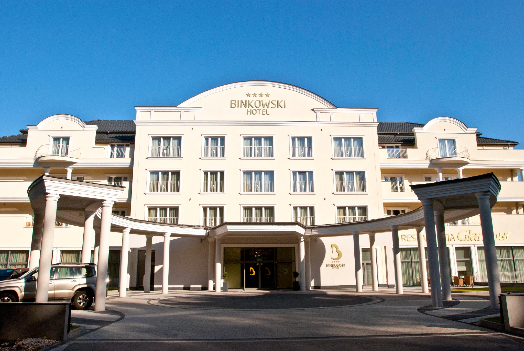 Elewacja hotelu Binkowski w klasycznym stylu z licznymi oknami z białymi ramami, kolumnami i podjazdem. Jasna fasada budynku na tle błękitnego nieba.