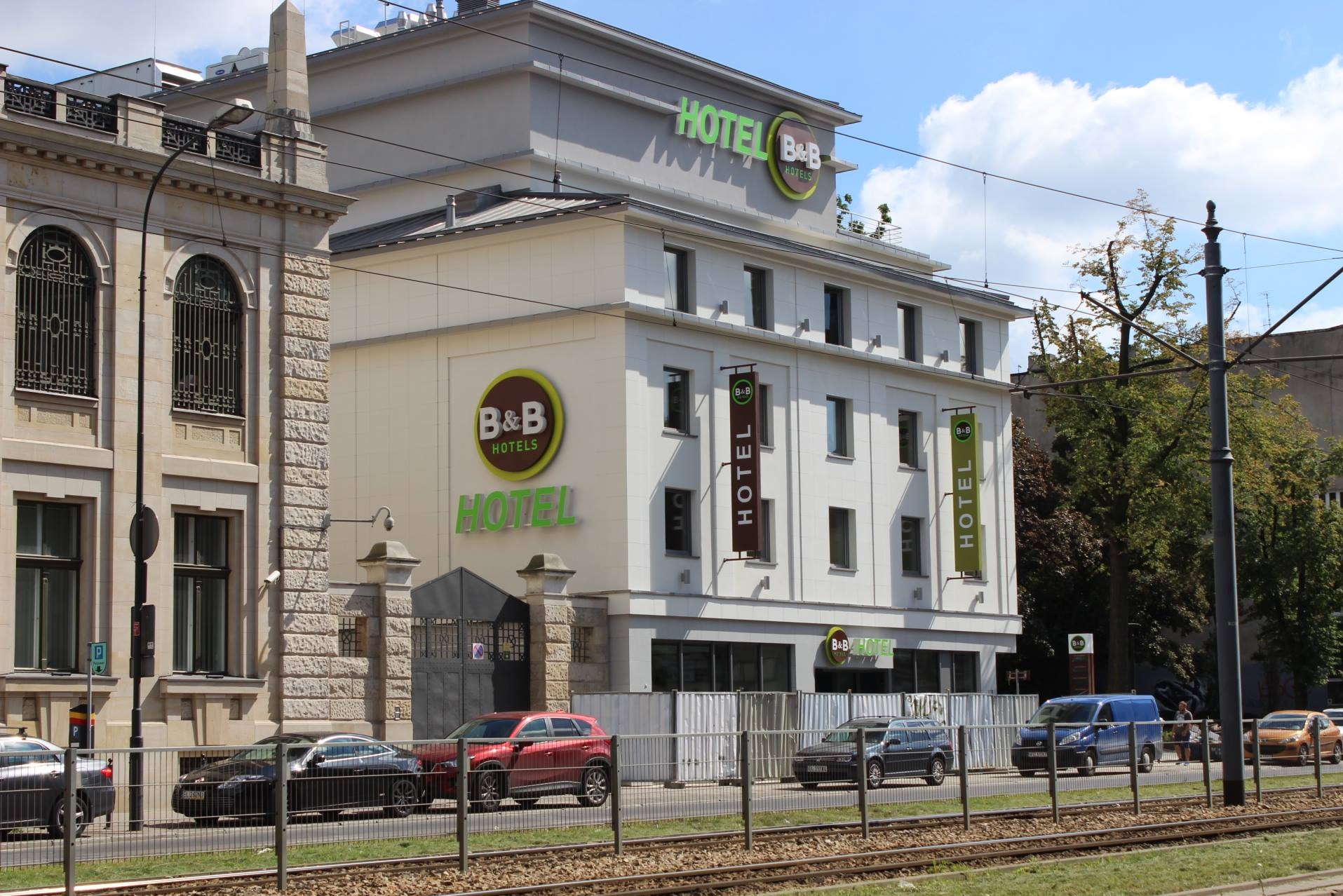 Elewacja hotelu B&B w jasnej kolorystyce z logo i zielonymi napisami. Widoczne okna, metalowe ogrodzenie i zaparkowane samochody. Ujęcie z poziomu ulicy.