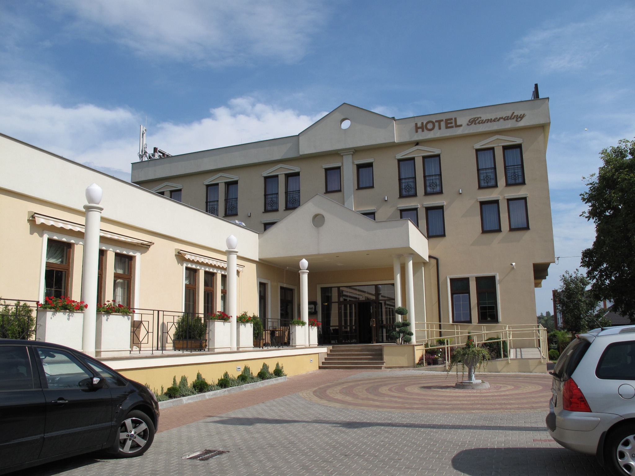 Elewacja trzypiętrowego hotelu z beżową fasadą, ciemnymi oknami i napisem 'Hotel Kameralny' pod błękitnym niebem. Widoczne auta na brukowanym parkingu.