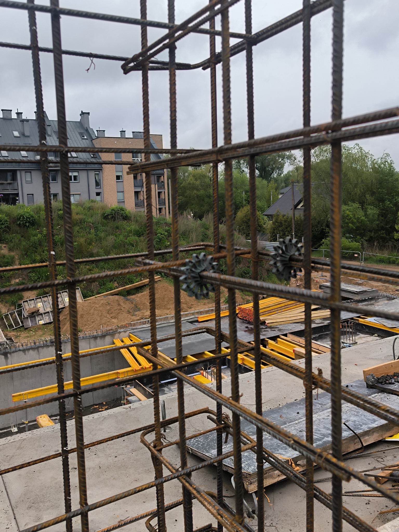 Siatka zbrojeniowa z dystansami na budowie, widok przez kratę na fundamenty, żółte belki i budynek w tle. Perspektywa z bliska, pochmurne niebo.