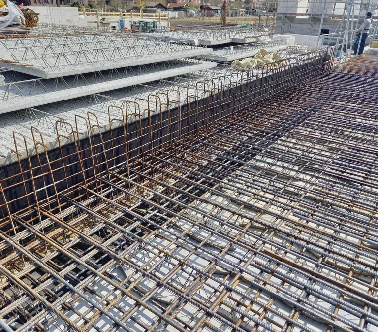 Siatka prętów na fundamencie z widocznymi elementami szalunku i częściowo ułożonymi prefabrykatami stropowymi. Ujęcie z góry, w świetle dziennym.