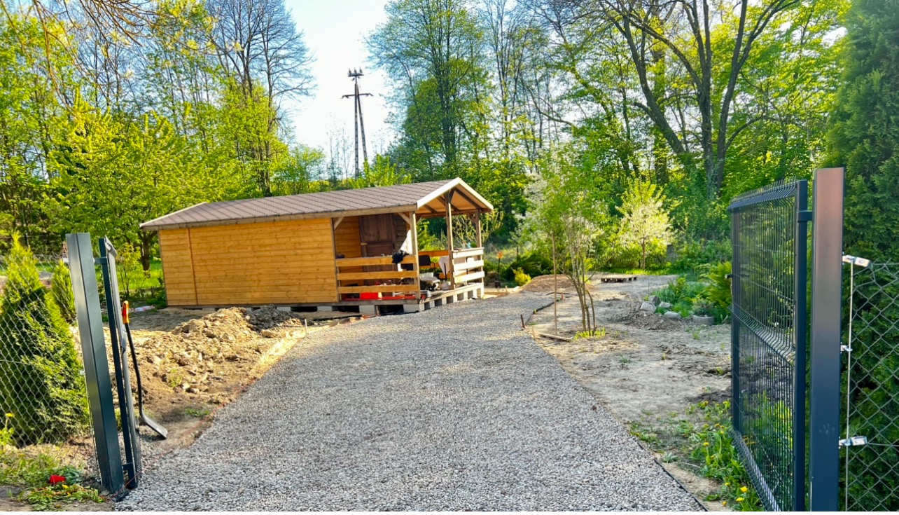 Utwardzona żwirem droga do drewnianej altany z brązowym dachem, widoczna przez otwartą, nowoczesną, metalową bramę w otoczeniu zieleni.