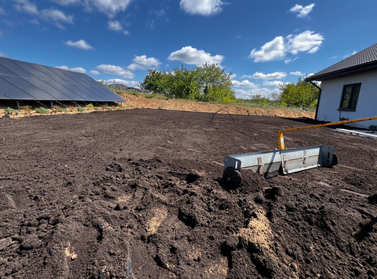 Niwelacja terenu pod budowę, widoczna spulchniona ziemia, narzędzie do wyrównywania, panele słoneczne w tle, dom z oknem, błękitne niebo.