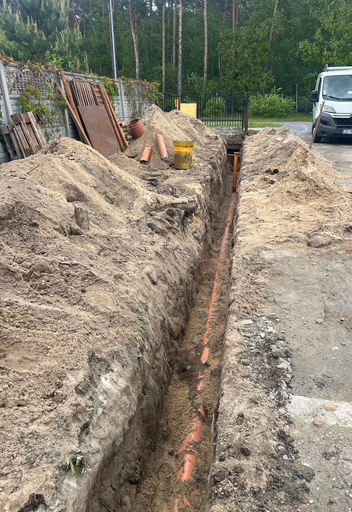 Wąski wykop z pomarańczową rurą na dnie, obok sterta ziemi, narzędzia i fragment vana. Prace ziemne w otoczeniu drzew i ogrodzenia.