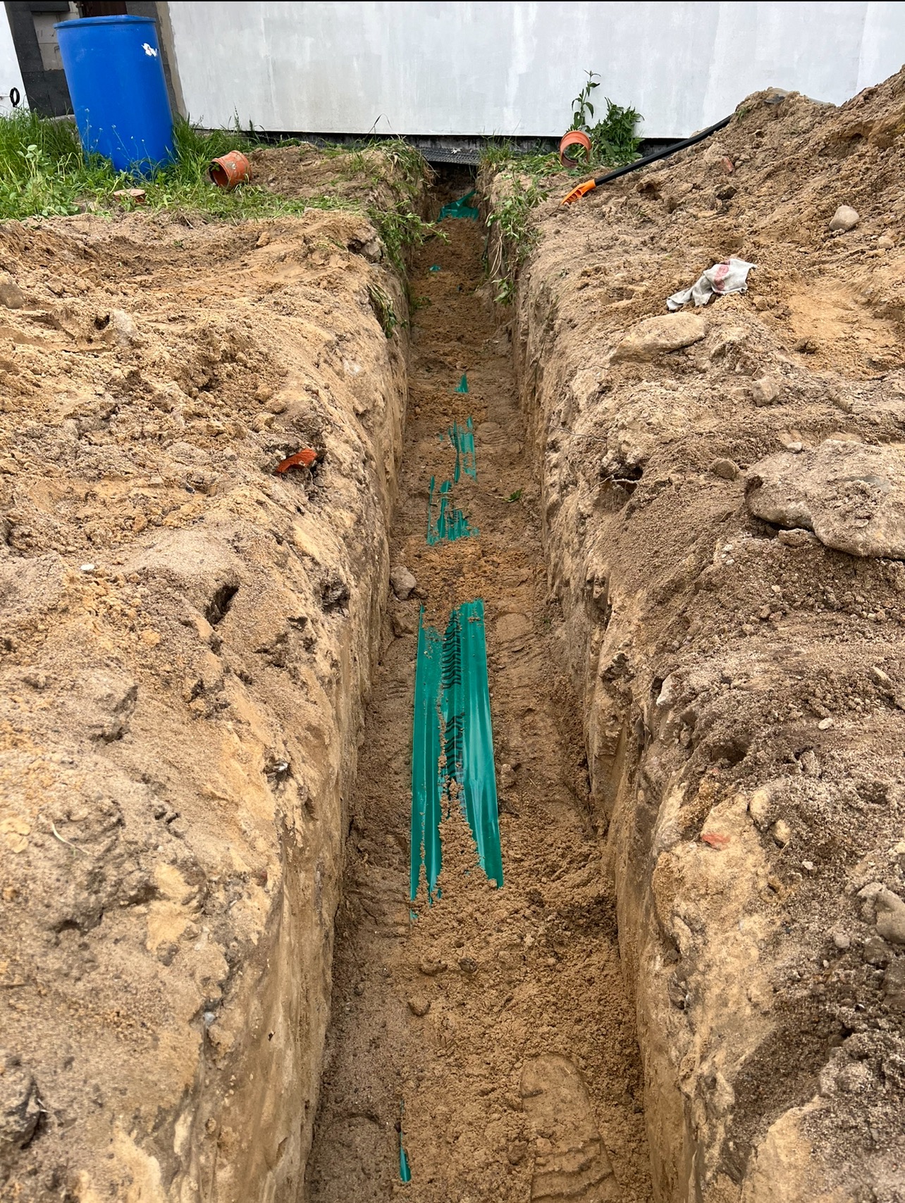 Wykopany rów z zielonymi rurami drenażowymi w ziemi, widok z góry. Na powierzchni widoczne narzędzia i niebieski pojemnik.