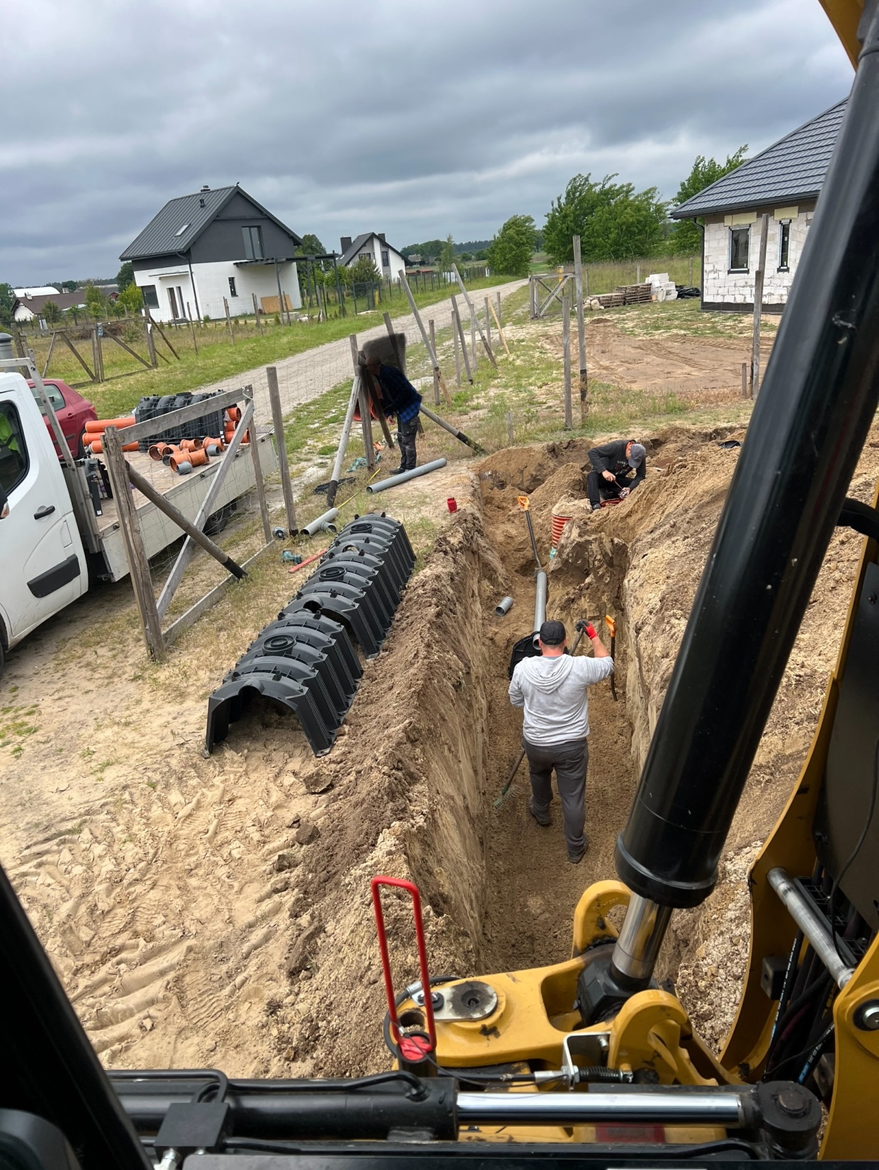 Prace ziemne: Wykop pod instalacje sanitarne z widocznymi pracownikami, rurami i elementami szalunku. Widok z kabiny koparki na plac budowy z domami w tle.
