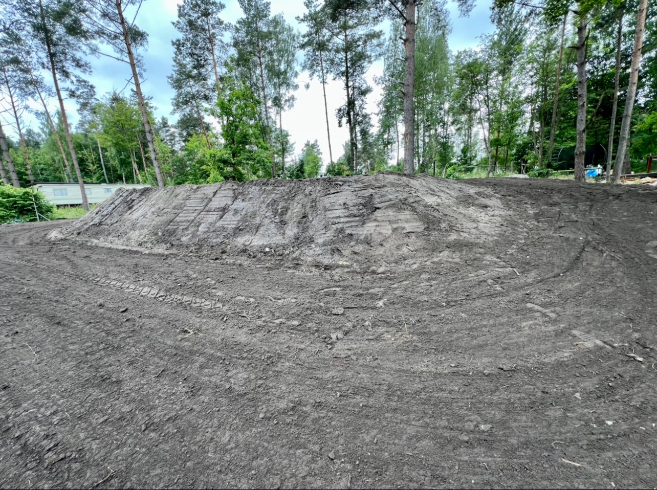 Uformowany nasyp ziemny z widocznymi śladami opon, otoczony zielenią drzew. Teren przygotowany, prawdopodobnie pod dalsze prace budowlane lub krajobrazowe.