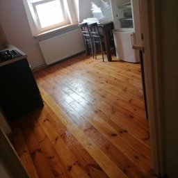 Usługi sprzątające Bombuś Marta Szczepaniak - Wnętrze mieszkania z drewnianą podłogą po sprzątaniu. Widoczne okno z łukiem, stół z krzesłami i lodówka. Podłoga czysta i błyszcząca.