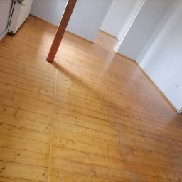 Usługi sprzątające Bombuś Marta Szczepaniak - Błyszcząca, odnowiona podłoga z desek w jasnym odcieniu drewna w pustym pokoju. Widoczny fragment ściany, drzwi i drewniany słup. Perspektywa z rogu pomieszczenia.