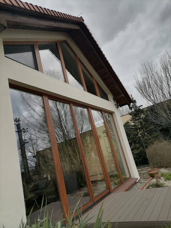 Duże okna z drewnianymi ramami w nowoczesnym domu, odbijające drzewa i pochmurne niebo. Widok z zewnątrz, z elementami ogrodu i drewnianym tarasem.