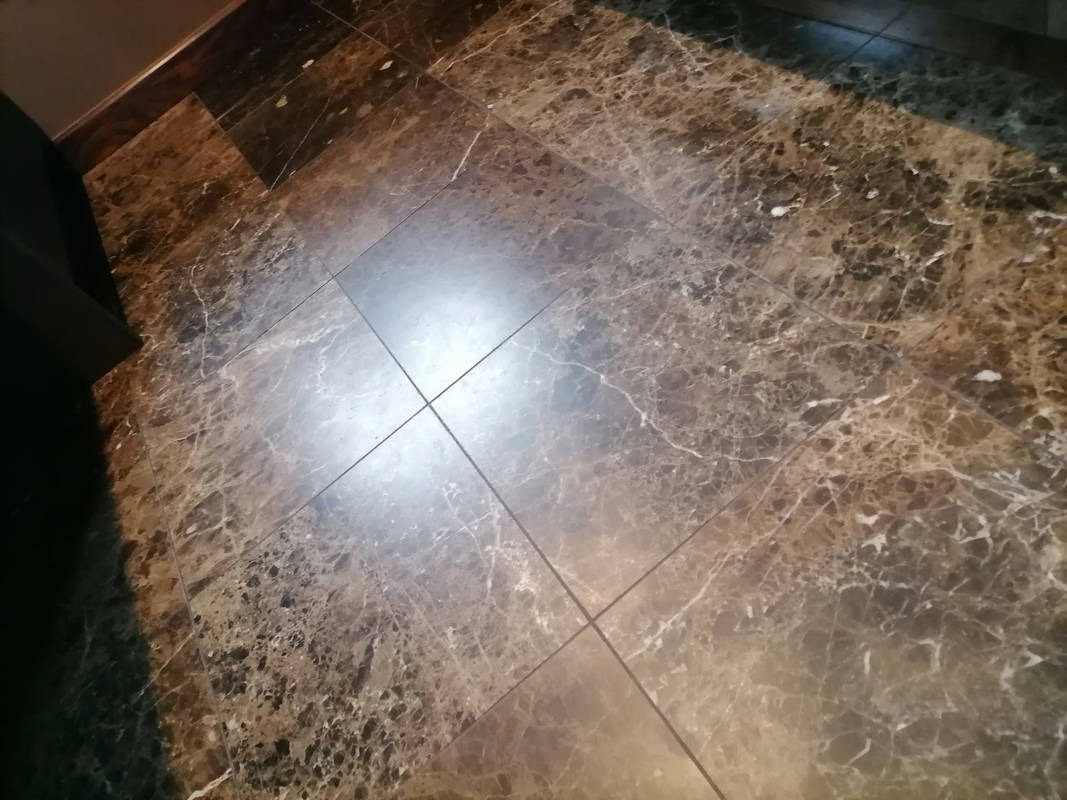 Błyszcząca, ciemnobrązowa podłoga z płytek imitujących marmur, z widocznymi fugami i odbiciem światła. W tle fragment mebla i listwa przypodłogowa.