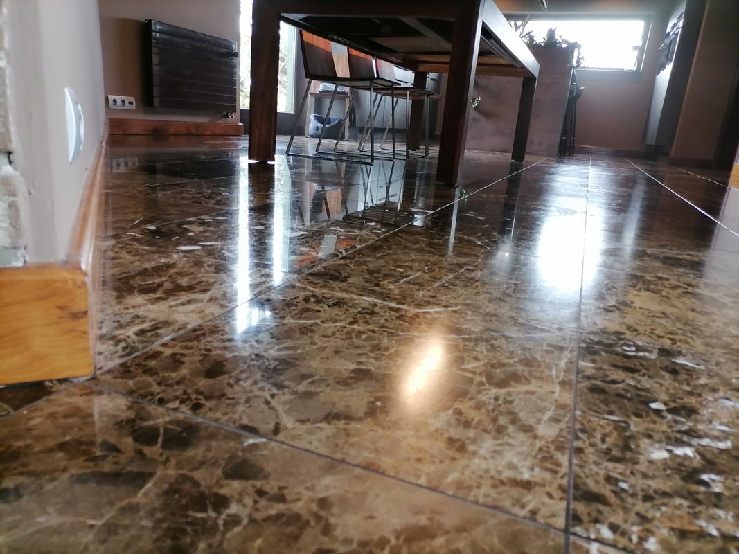 Błyszcząca, brązowa podłoga z płytek imitujących marmur, odbijająca światło. W tle fragment stołu i krzesła w jasnym wnętrzu.