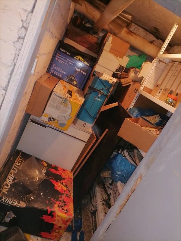 Zagracone pomieszczenie ze stertą pudeł, sprzętów i śmieci pod ścianą. Widoczne regały z dokumentami. Perspektywa z góry.