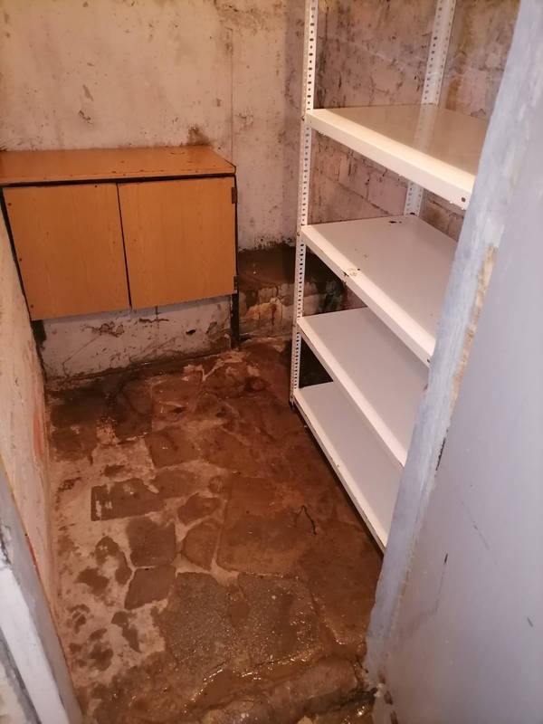 Zaniedbane pomieszczenie z mokrą, kamienną podłogą, szafką i metalowym regałem. Brudne ściany z zaciekami i odpadającym tynkiem. Widoczny brak dbałości o czystość.