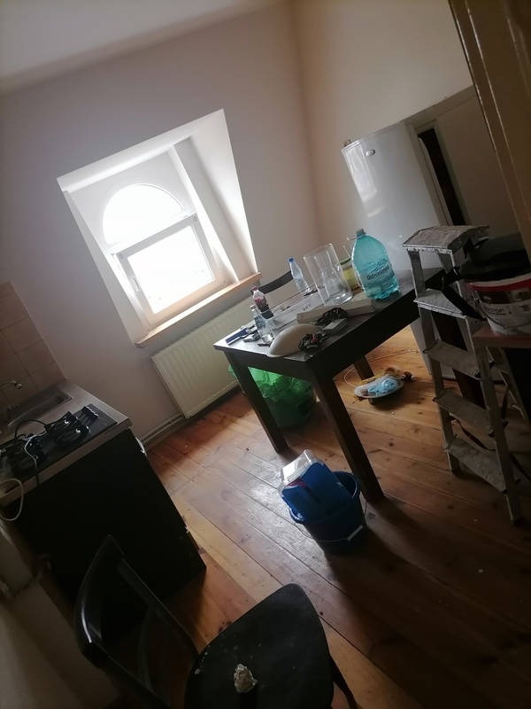 Pokój przed sprzątaniem: bałagan na stole, podłodze i wokół kuchenki. Widoczne ślady użytkowania i zaniedbania. Przygotowanie do czyszczenia.
