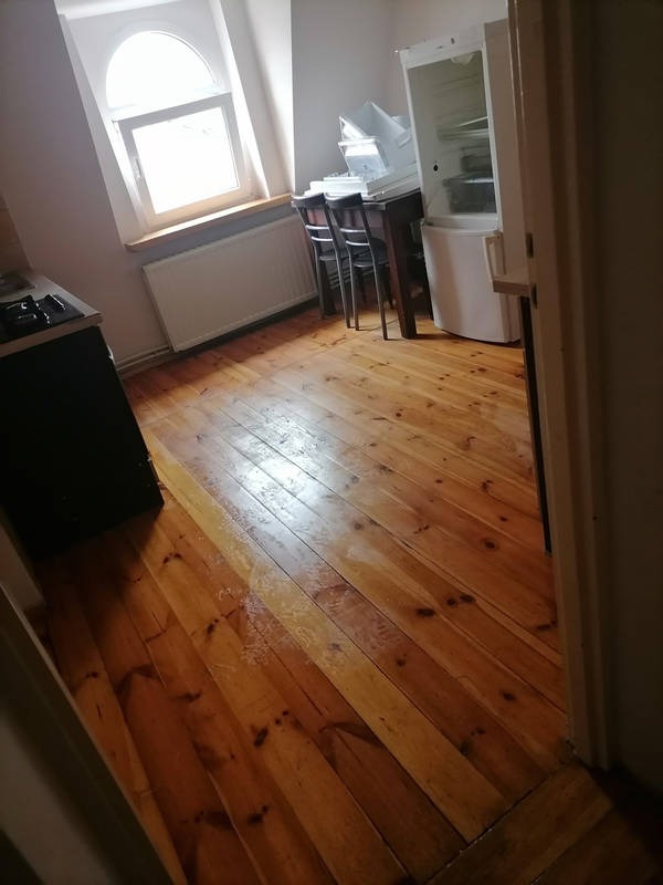 Wnętrze mieszkania z drewnianą podłogą po sprzątaniu. Widoczne okno z łukiem, stół z krzesłami i lodówka. Podłoga czysta i błyszcząca.