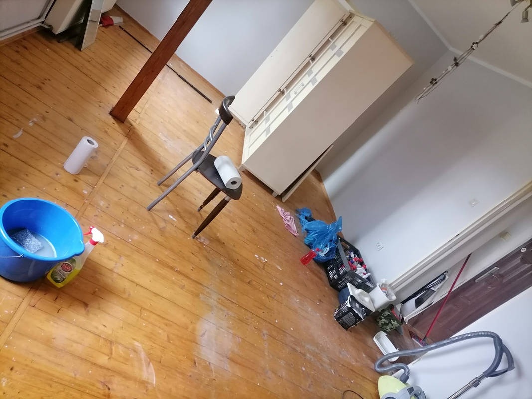 Podłoga po remoncie z wiadrem, środkiem czyszczącym, krzesłem i rolką papieru. Widoczne ślady farby i zabrudzenia. Sprzątanie po remoncie w trakcie.