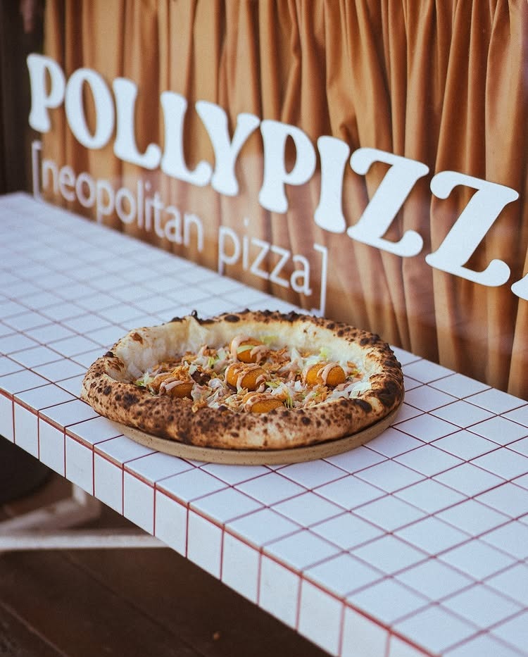 Neapolitańska pizza z dodatkami na tle logotypu 'Polly Pizz'. Pizza na białym stole z czerwoną kratą, brązowe zasłony w tle. Jasne, naturalne światło.