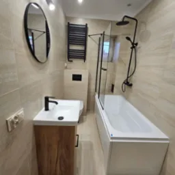 Wykończona łazienka w beżowych płytkach z drewnianą szafką pod umywalką, czarną armaturą i grzejnikiem. Kabina prysznicowa z czarnymi ramami i szklanymi drzwiami.