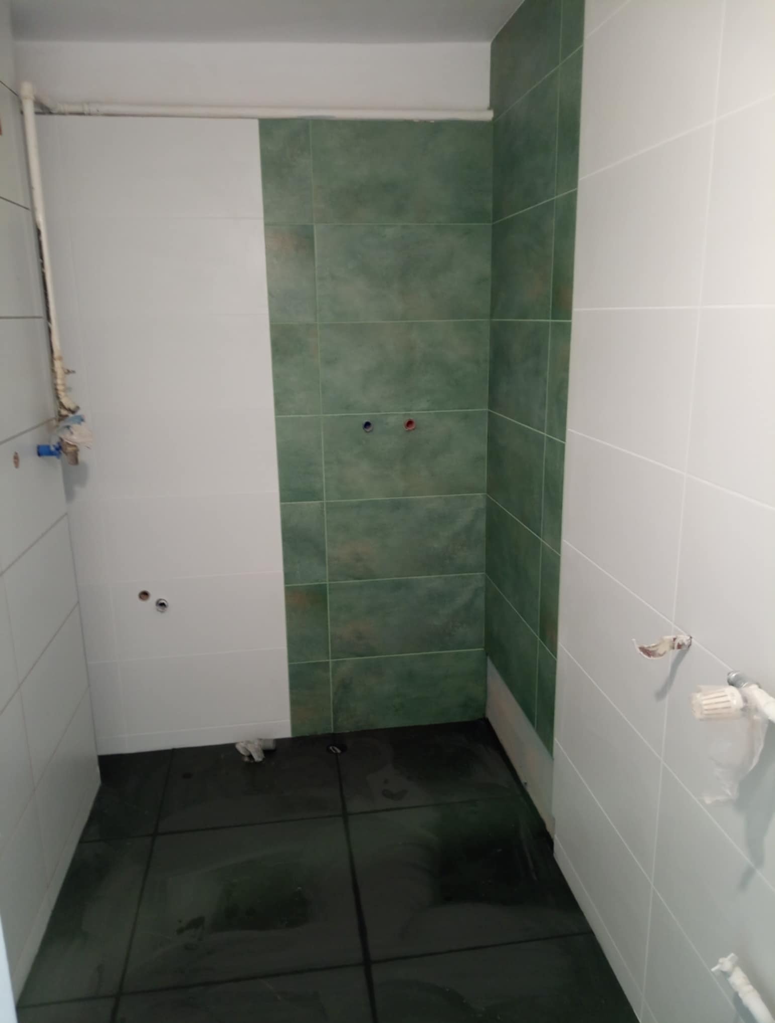 Wnętrze łazienki w trakcie prac wykończeniowych: białe i zielone płytki na ścianach, ciemna podłoga, widoczne instalacje wodne. Częściowo wykończone pomieszczenie.