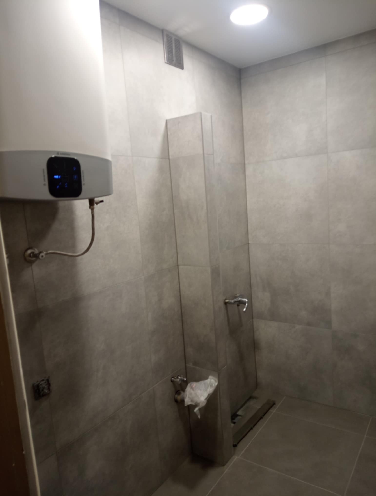 Wykończona łazienka z szarymi płytkami, widoczny bojler elektryczny, bateria i odpływ liniowy. Minimalistyczny design, neutralna kolorystyka.