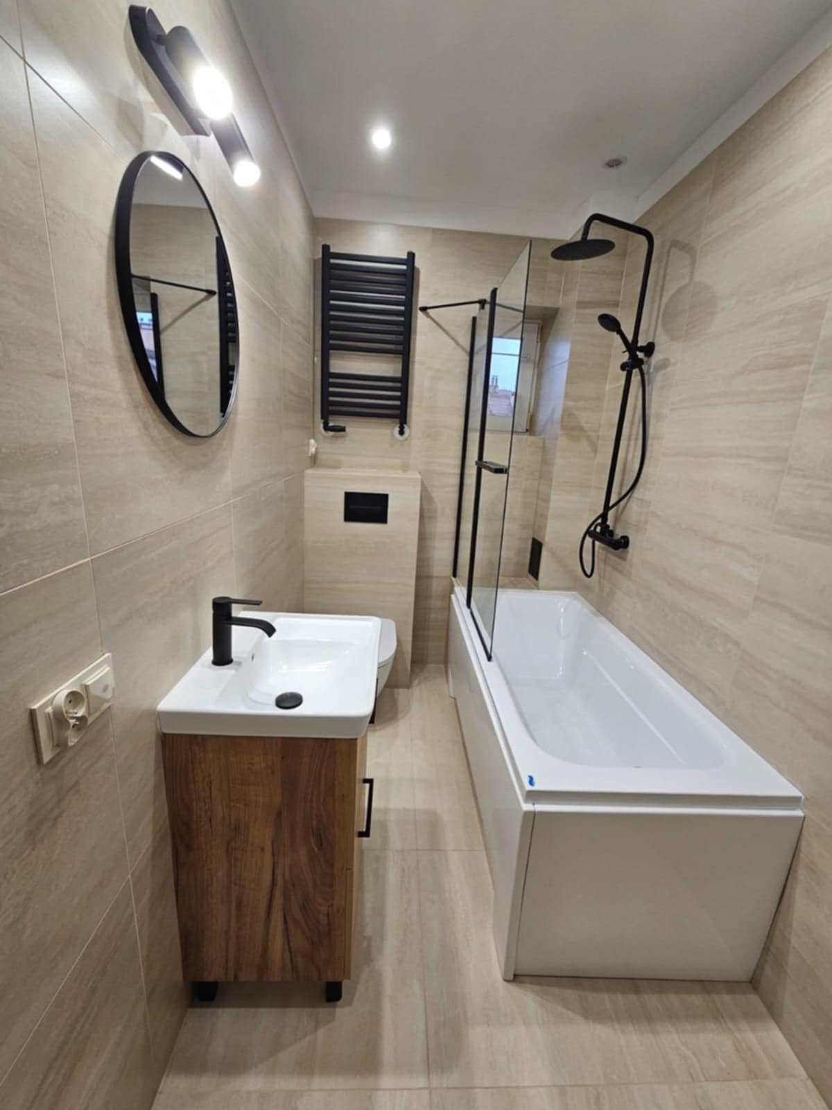 Wykończona łazienka w beżowych płytkach z drewnianą szafką pod umywalką, czarną armaturą i grzejnikiem. Kabina prysznicowa z czarnymi ramami i szklanymi drzwiami.
