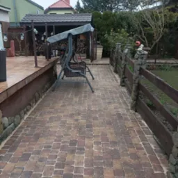 Brązowa kostka brukowa na podwórku z drewnianym ogrodzeniem i huśtawką. W tle drewutnia i zieleń. Widok na fragment podwórka.