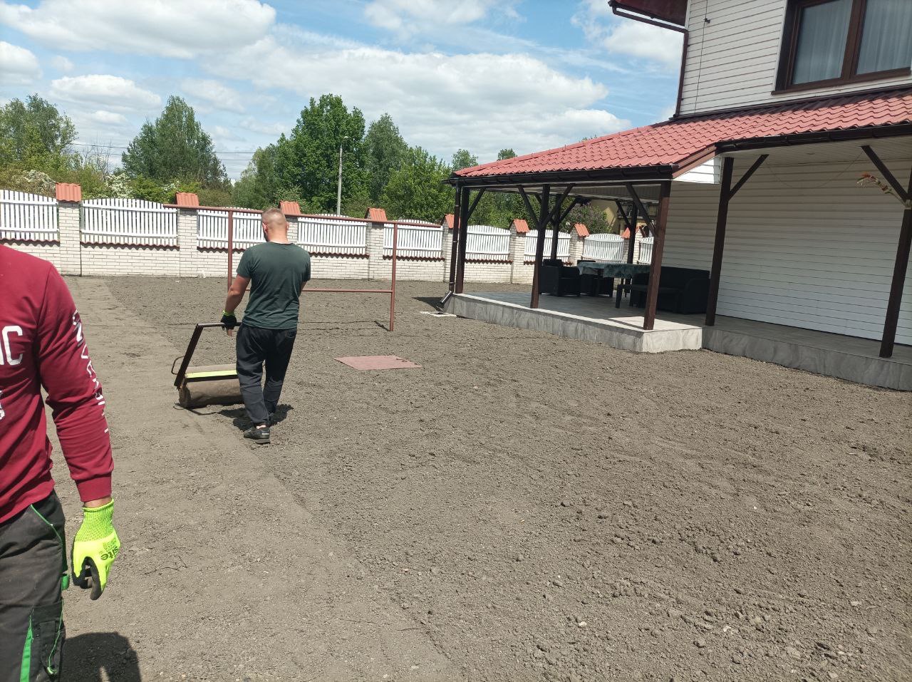Przygotowanie gruntu pod trawnik: mężczyzna używa walca na nierównym terenie, w tle białe ogrodzenie i altana z czerwonym dachem. Prace ziemne.