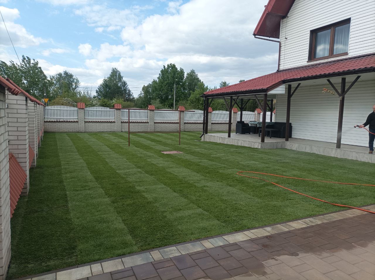 Świeżo skoszony trawnik z wyraźnymi pasami, otoczony białym ogrodzeniem i domem z czerwonym dachem. Osoba podlewa trawnik w prawym rogu.