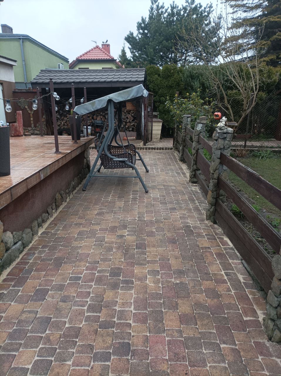 Brązowa kostka brukowa na podwórku z drewnianym ogrodzeniem i huśtawką. W tle drewutnia i zieleń. Widok na fragment podwórka.