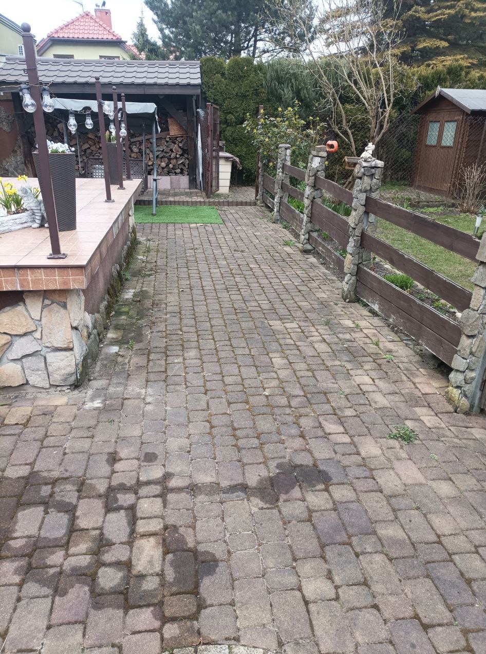 Ułożona kostka brukowa na podwórku z drewniano-kamiennym ogrodzeniem i elementami architektury ogrodowej w tle, widok z perspektywy.