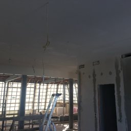 Braim Company Sp z o.o - Wnętrze w trakcie prac wykończeniowych z widocznymi przewodami elektrycznymi zwisającymi z sufitu i drabiną w tle. Surowe ściany i duże okna.