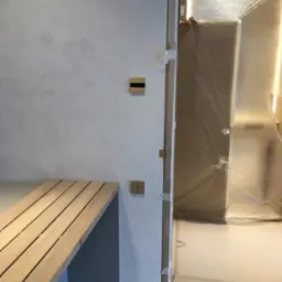 Minimalistyczne włączniki światła na teksturowanej ścianie, drewniana ławka, zabezpieczone folią drzwi. Detal wykończenia wnętrza w trakcie prac.