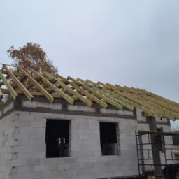 Wznoszenie domu z białych bloczków: ściany z otworami okiennymi i drewniana konstrukcja dachu w trakcie budowy. Jesienna sceneria, pochmurne niebo.