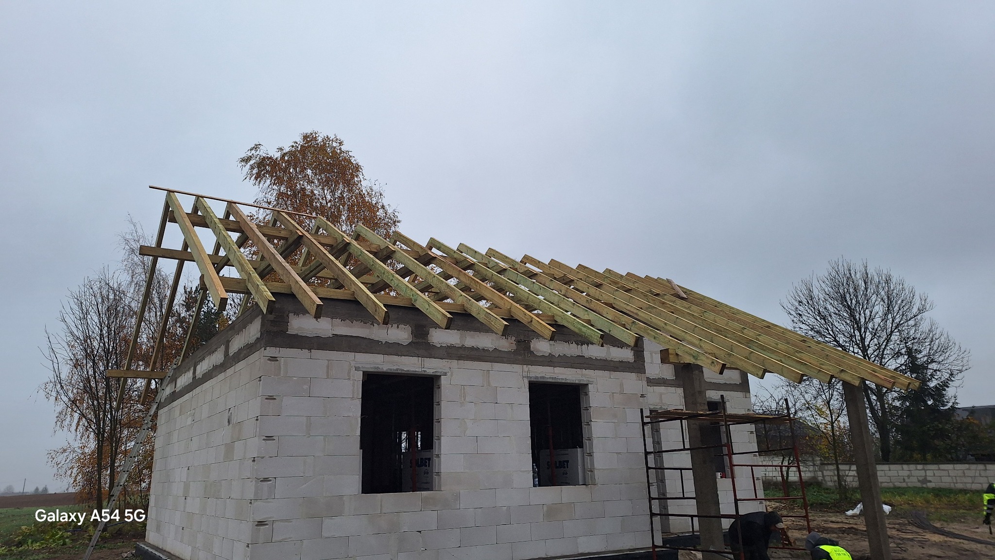 Wznoszenie domu z białych bloczków: ściany z otworami okiennymi i drewniana konstrukcja dachu w trakcie budowy. Jesienna sceneria, pochmurne niebo.