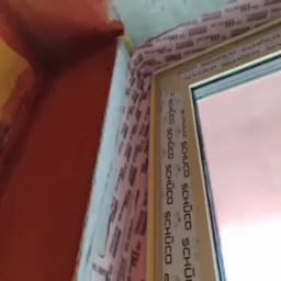 Szczegółowe ujęcie okna Schüco z uszczelnieniami Soudal, pomarańczowe tło. Kątowe ujęcie podkreśla precyzję montażu i dbałość o detale wykończenia.